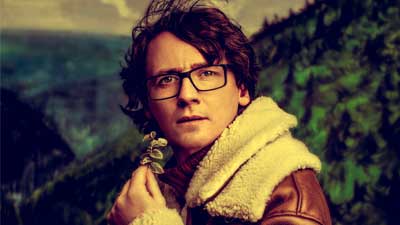 Comedian Ed Byrne wearing a fur lined coat, looking straight at the camera 