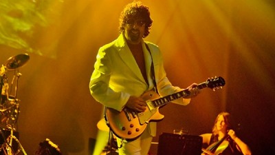 A band member of the ELO Experience holds a guitar in front of a deep orange background. 