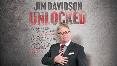 Comedian Jim Davidson stands infront of a grey wall with the text 'Jim Davidson Unlocked'