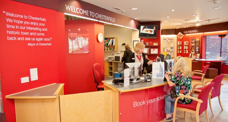 A brightly coloured photograph of the Visitor Information Centre. The walls are red and there is a light oak wooden counter. The cashier is stood behind the desk and is serving a customer who is sat down on a chair. 