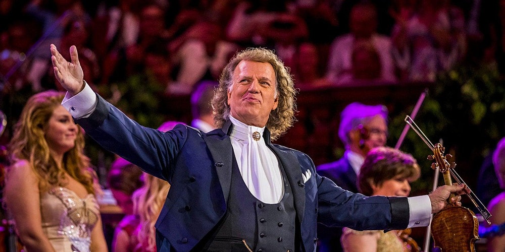Andre Rieu stands on stage with his arms outstretched. 