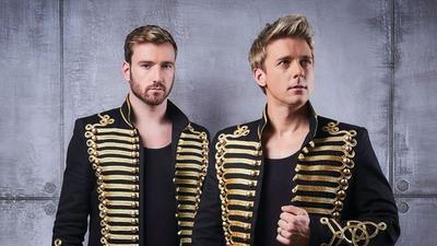 Singers Jonathon Ansell and Jai McDowall stand against a grey backdrop. 