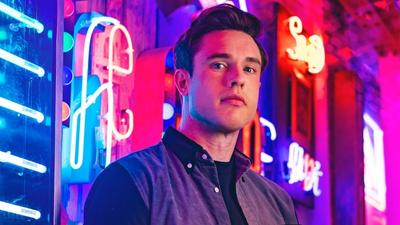 Comedian Ed Gamble stands against a neon lit background. 