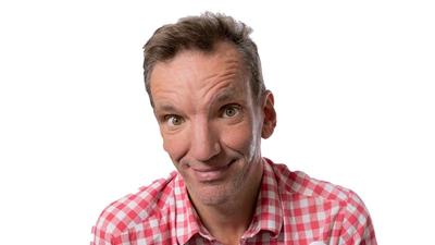 A headshot of comedian Henning Wehn wearing a red and white checked shirt.