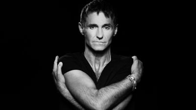 A black and white image of singer Marti Pellow against a black backdrop with his arms wrapped around himself. 