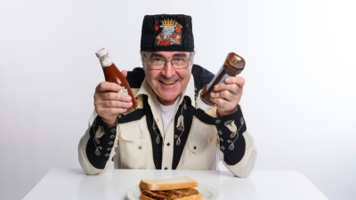 Comedian Danny Baker sits at a table holding with a sausage sandwich in front of him. He has red sauce in one hand and brown sauce in the other. 