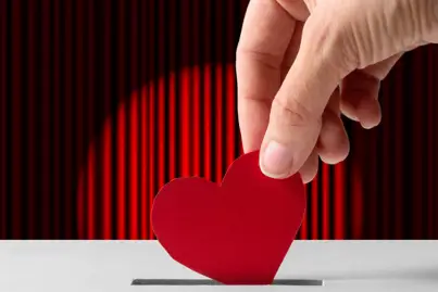 A photograph of a stage curtain. A large hand is coming above teh stage and posting a red paper heart into a donations box.