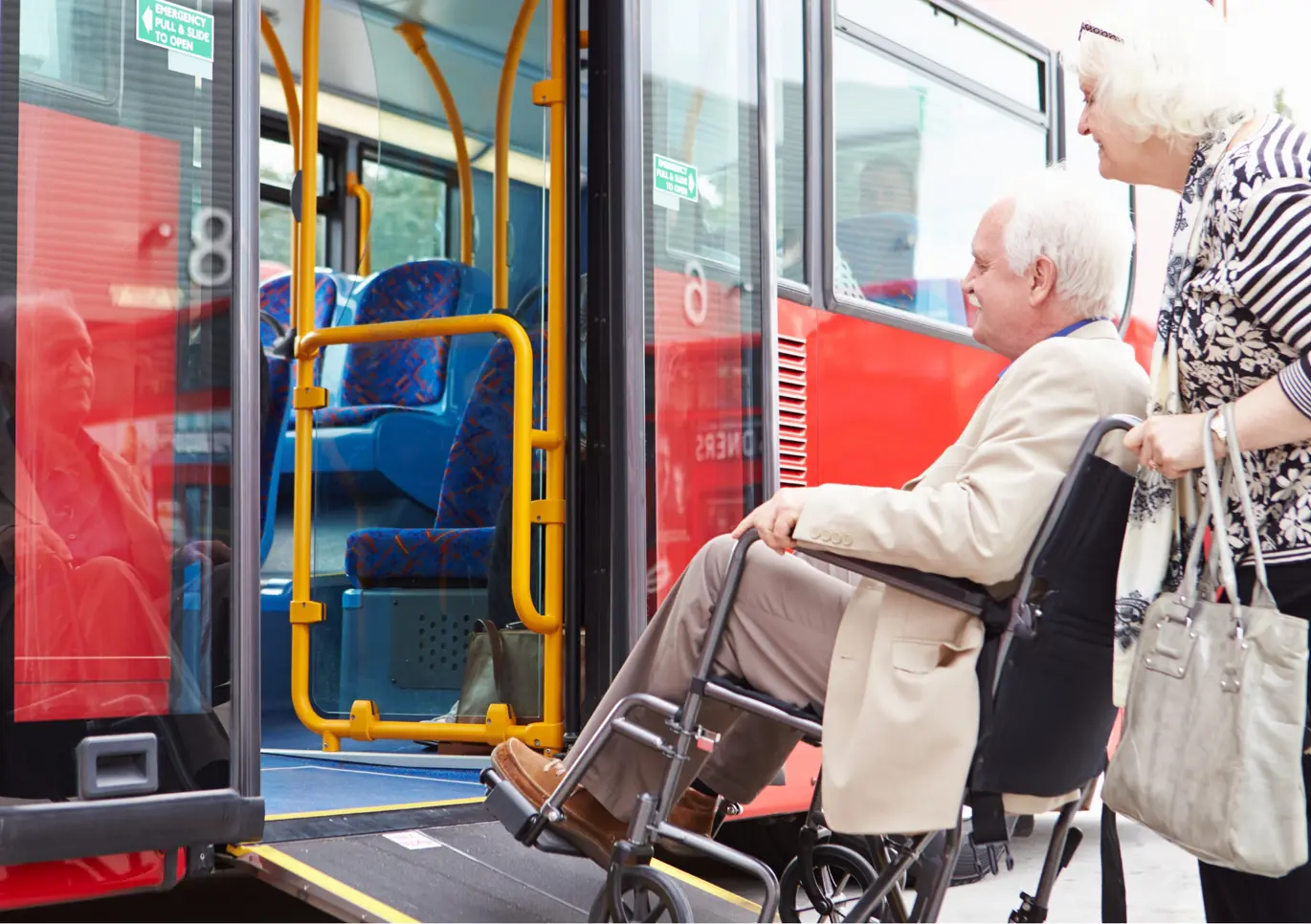 Wheelchair user using a ramp to get onto a red public transport bus. The man with white hair is being pushed up the ramp by a lady with white hair.