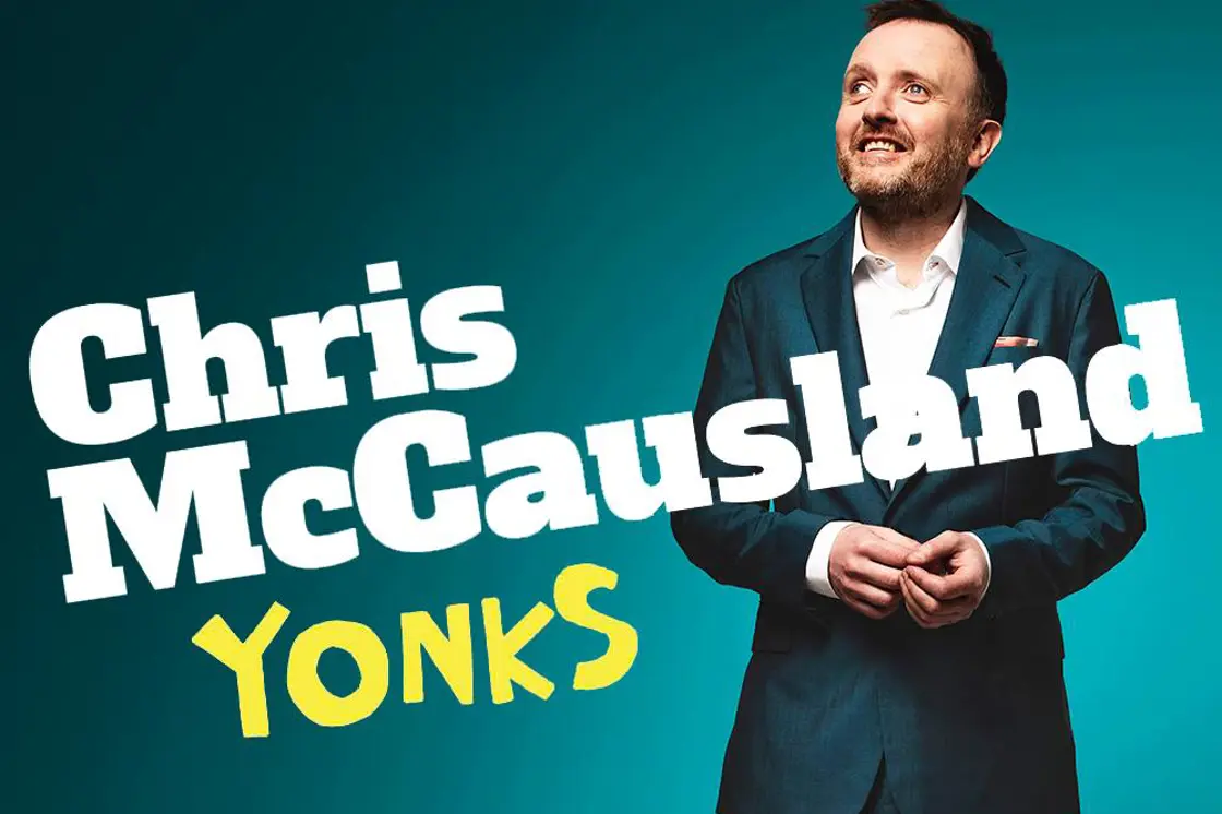 Comedian Chris McCausland wears a suit as he stands against a turquoise background.