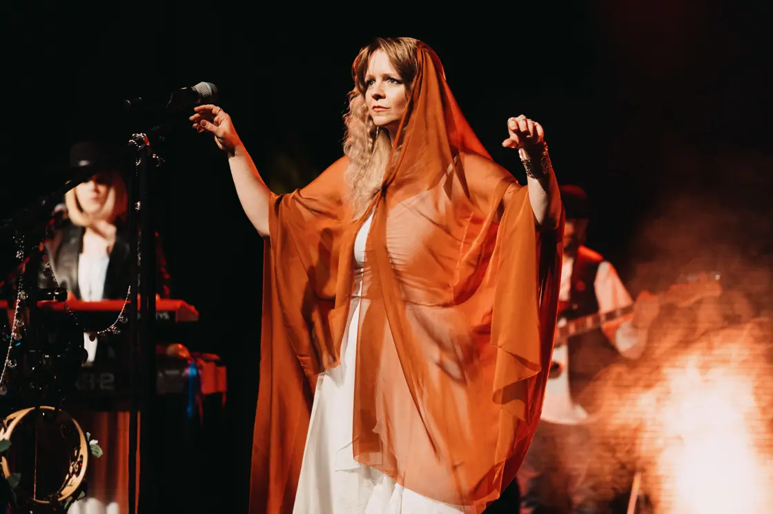 A member of The Fleetwood Mac Legacy wears a red floaty cape as she stands on stage. 
